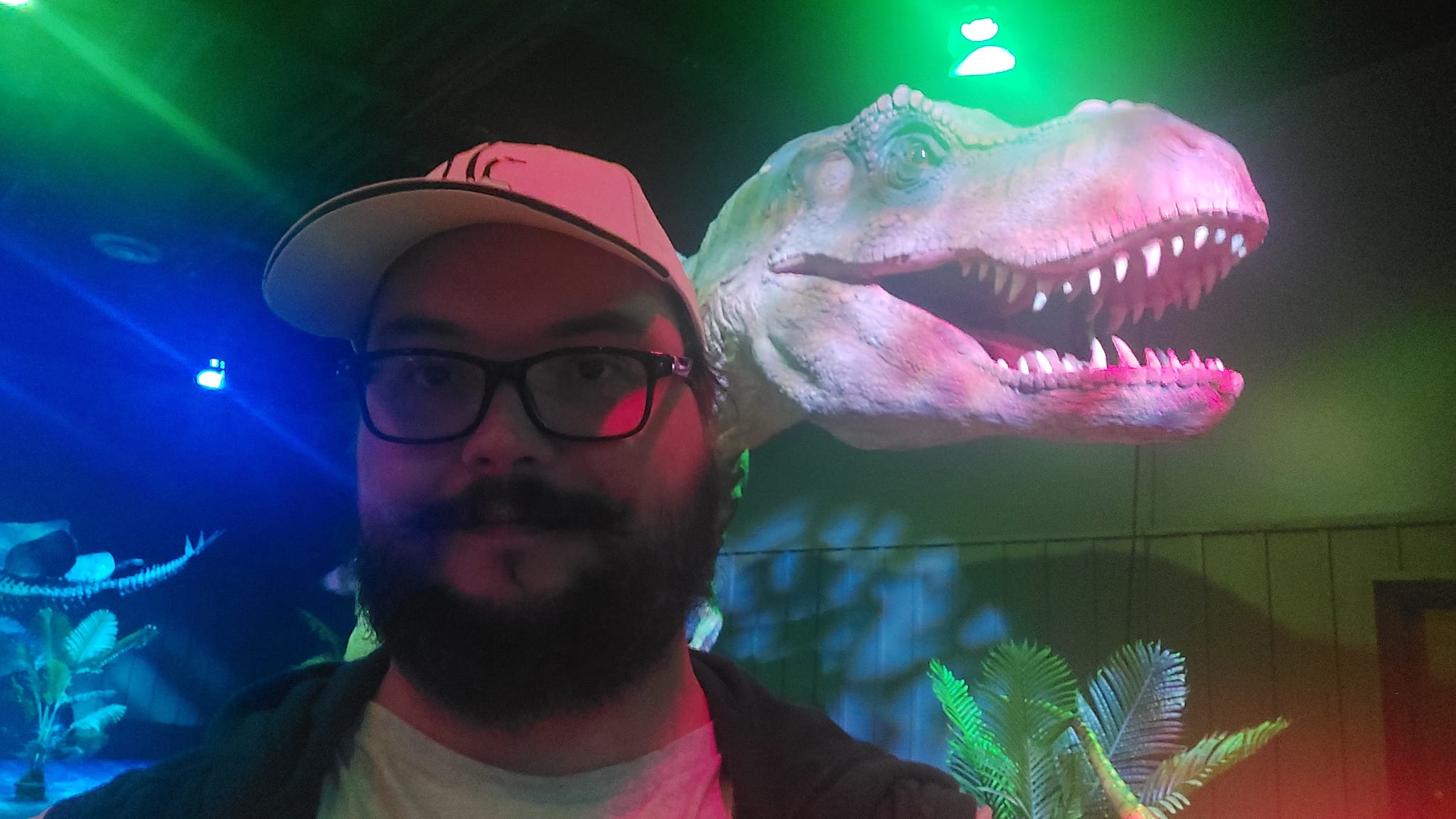 A man with a ball cap and bushy facial hair is standing in front of a tyranosaurus animatronic